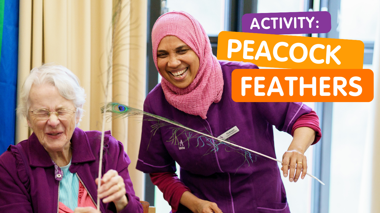 A South-Asian female carer plays with peacock feathers with a white, female care home resident. They both laugh